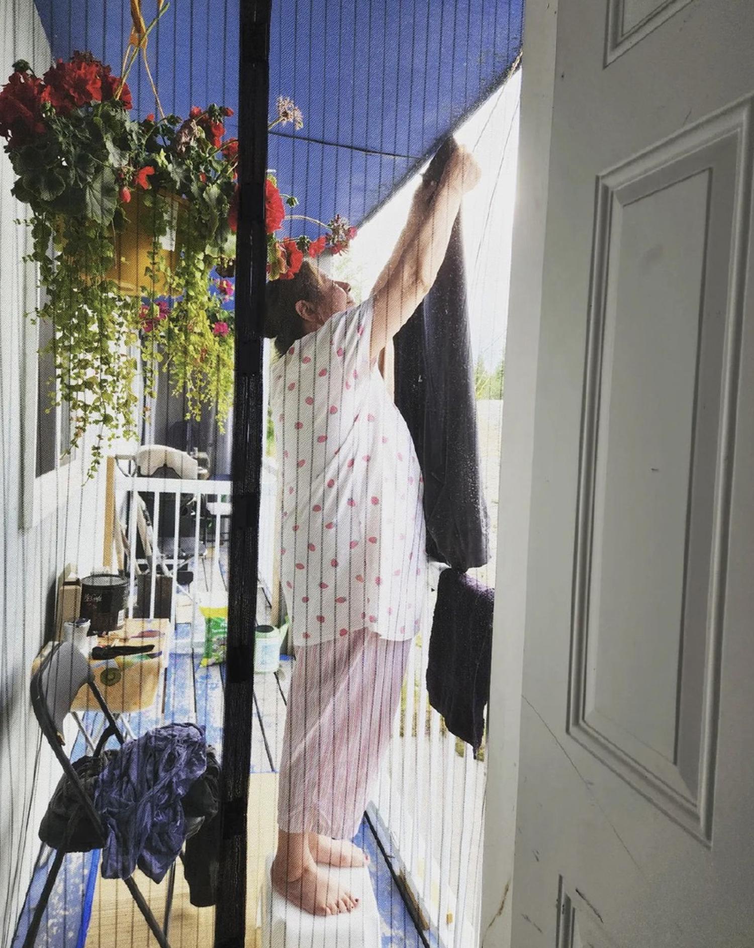 Image of woman hanging laundry on a porch taken through a doorway with flowers in the foreground.