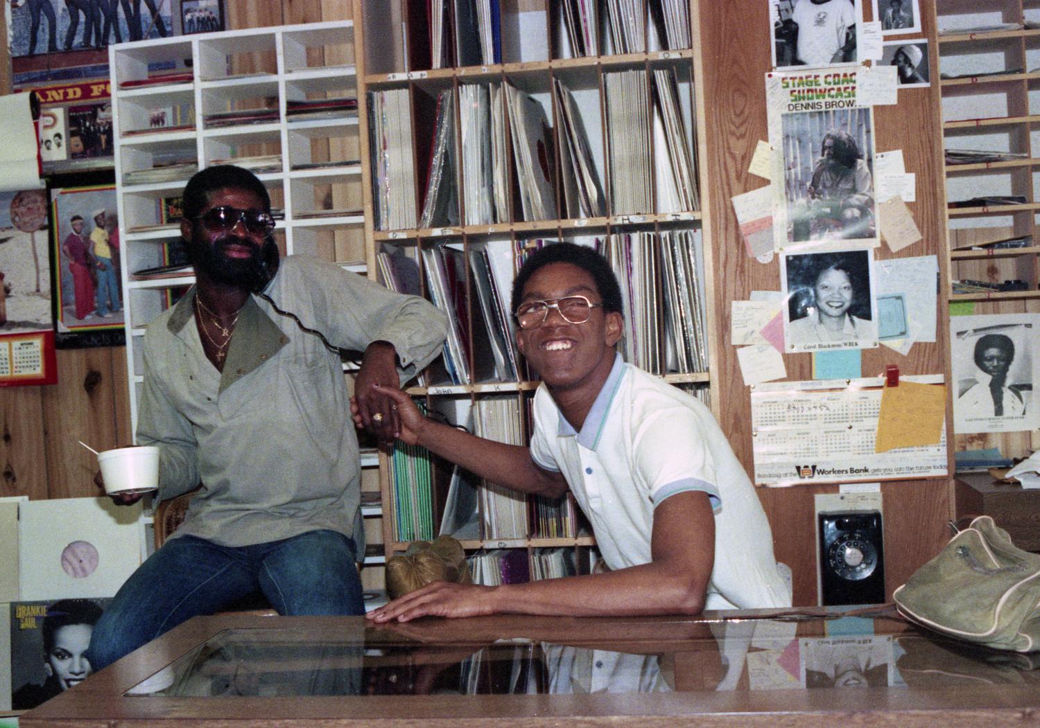 Two men seated, one on the phone eating and shaking the hand of the other.