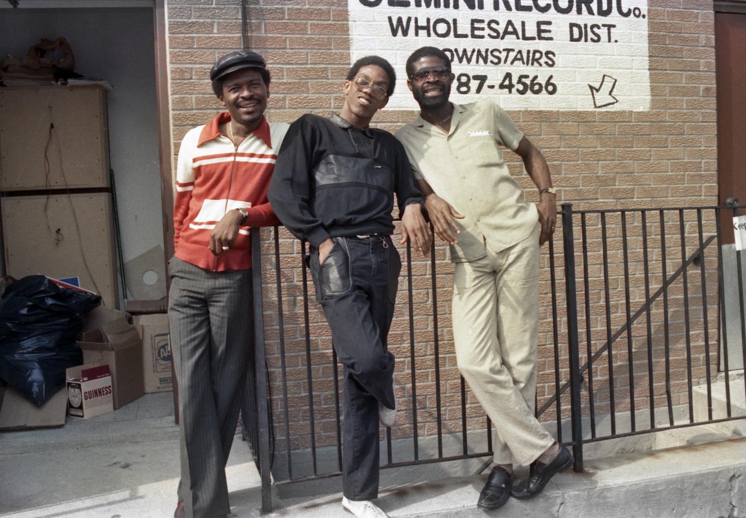 Three men leaning against a railing outside of a building with a sign reading Gemini Records Co.
